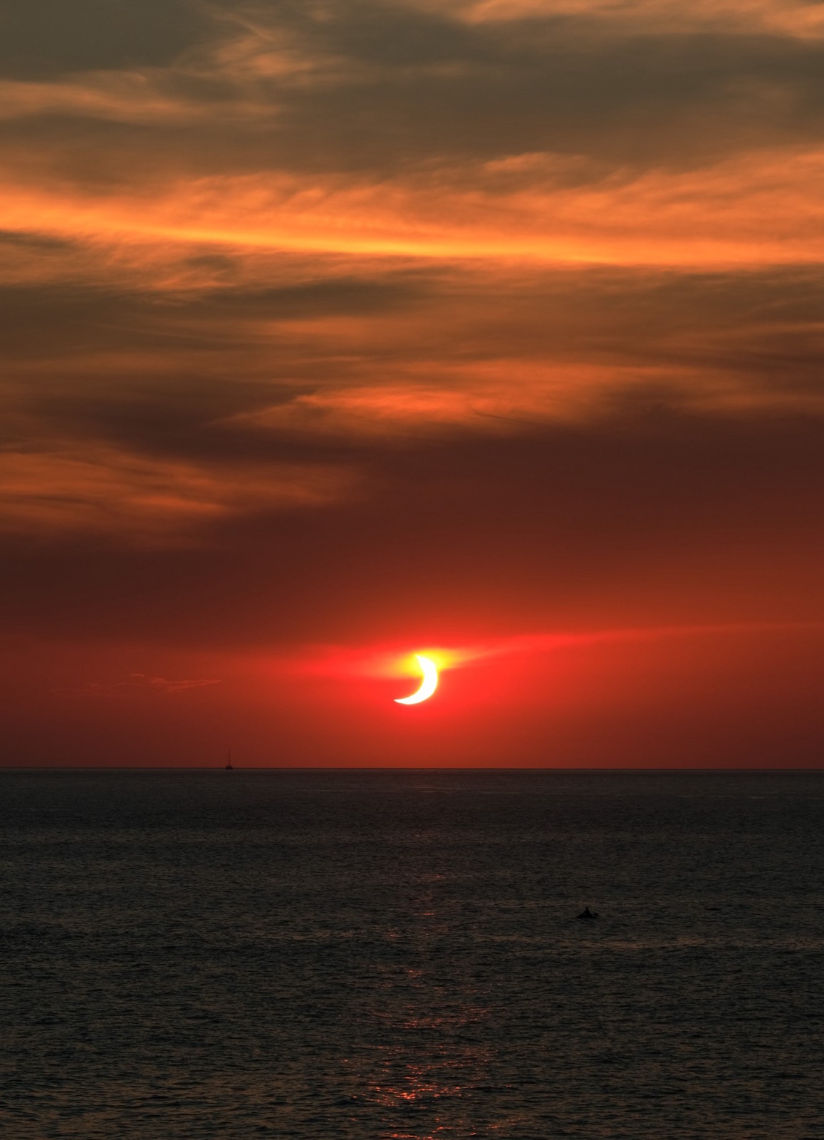 A partially eclipsed sun setting over the sea in an orange sky.