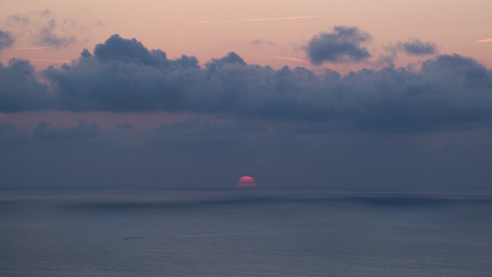 A red sun sinking into a calm sea under a band of clouds.